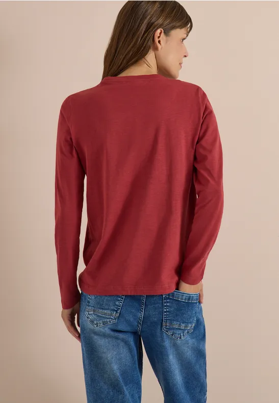 Frau steht mit dem Rücken zur Kamera, trägt rotes Langarmshirt und blaue Jeans.