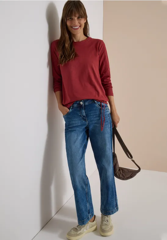 Junge Frau lehnt an der Wand, trägt rotes Oberteil, blaue Jeans und eine braune Umhängetasche.