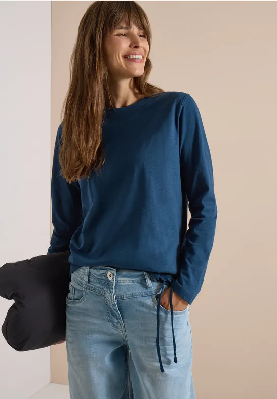 Eine Frau in blauem Langarmshirt und Jeans lehnt lächelnd an einer Wand, hält ein Kissen in der Hand.