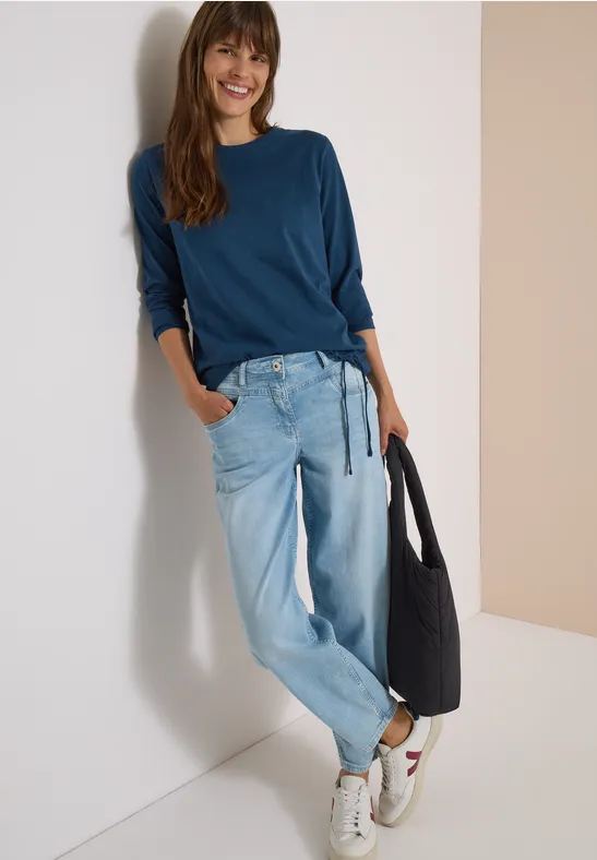 Frau lehnt an Wand, trägt blaues Shirt, helle Baggy-Jeans, weiße Sneaker und hält schwarze Tasche.