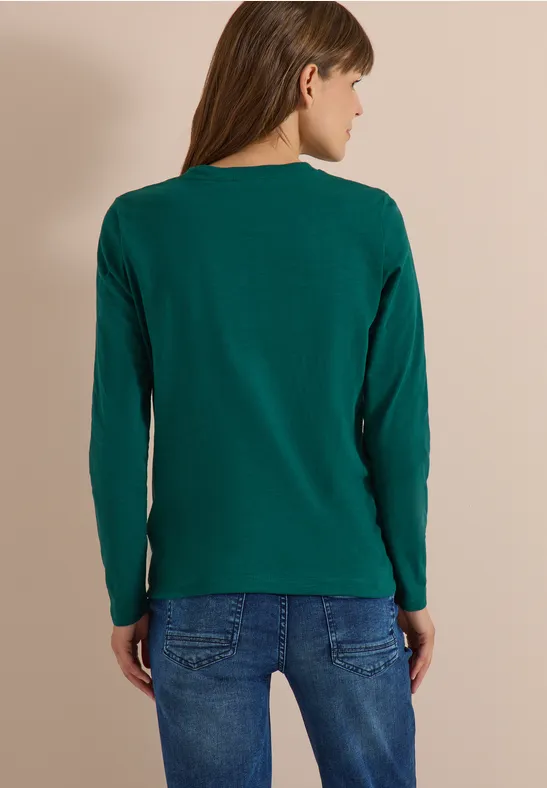 Frau von hinten trägt grünes Langarmshirt und blaue Jeans vor beigem Hintergrund