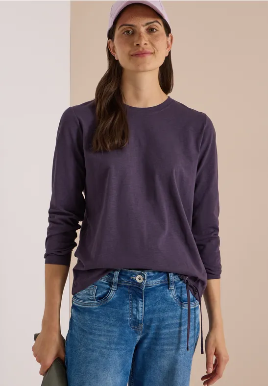 Frau in lila Langarmshirt, blauer Jeans und Cap vor neutralem Hintergrund.