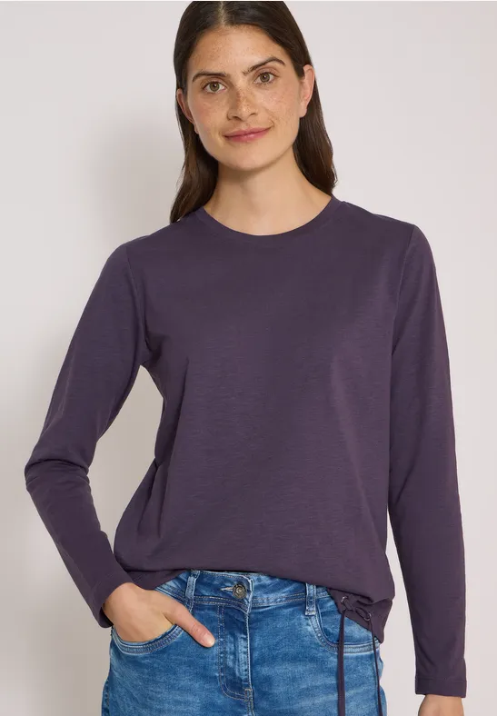 Eine Frau in dunkelviolettem Langarmshirt und Jeans vor hellem Hintergrund.