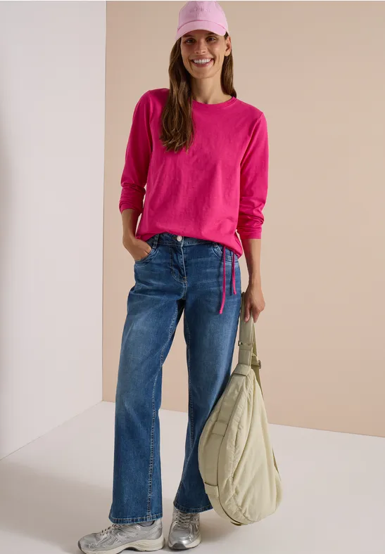 Frau in pinkem Langarmshirt, blauen Jeans, rosa Cap und großer Schultertasche