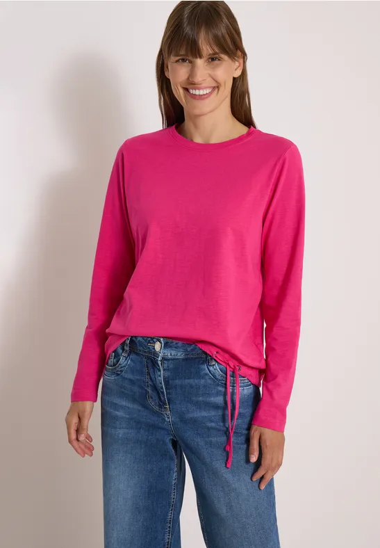 Frau steht vor hellem Hintergrund, trägt pinken Pullover und blaue Jeans und lächelt in die Kamera.