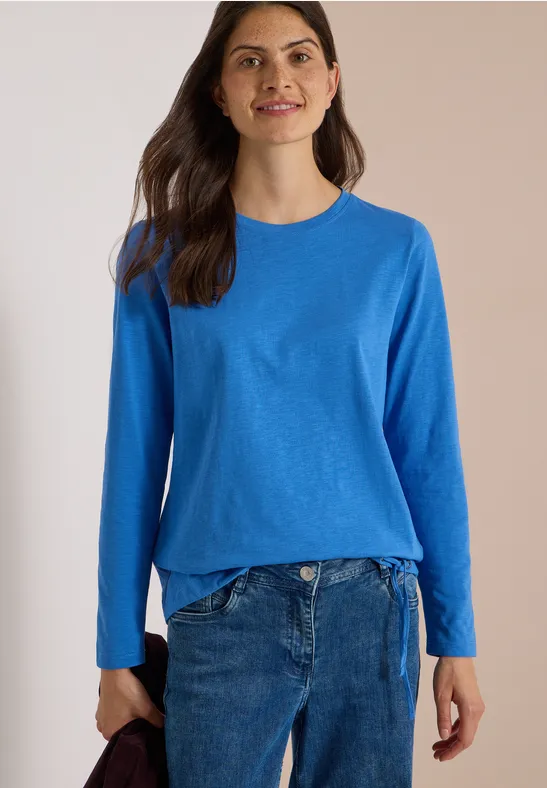 Eine Frau steht vor hellem Hintergrund, trägt einen hellblauen Pullover und Jeans und lächelt.