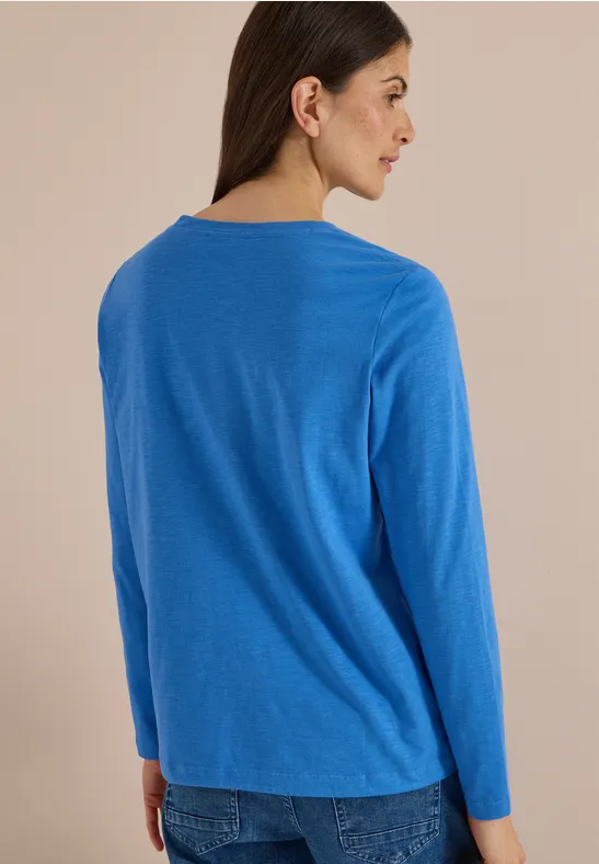 Frau von hinten in blauem Langarm-Pullover und Jeans vor neutralem Hintergrund.