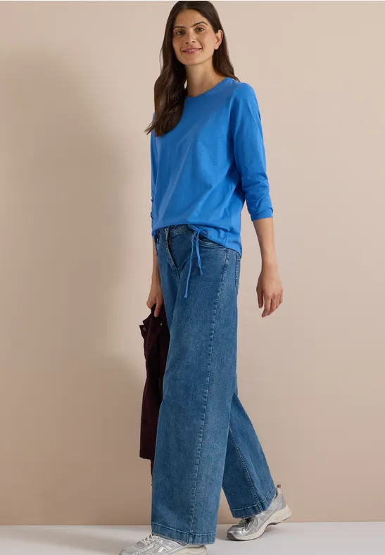 Frau in blauem Langarmshirt und weitem Jeansrock mit Taillenschnürung, weiße Sneaker, hält dunkle Tasche.