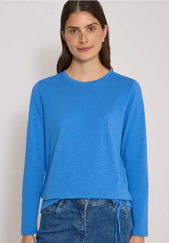 Frau im blauen Langarmshirt und Jeans vor hellem Hintergrund.