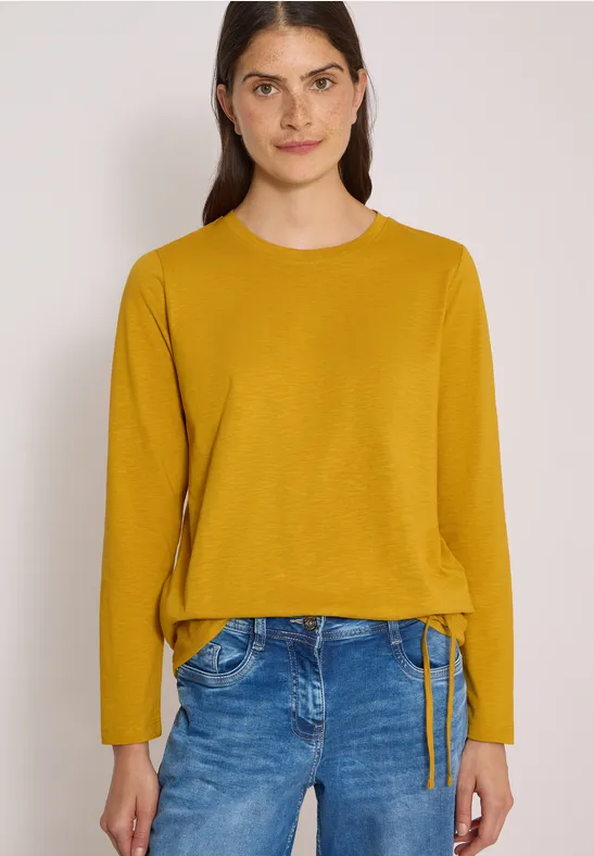 Frau mit dunklen Haaren trägt einen senfgelben Pullover und Jeans vor neutralem Hintergrund.