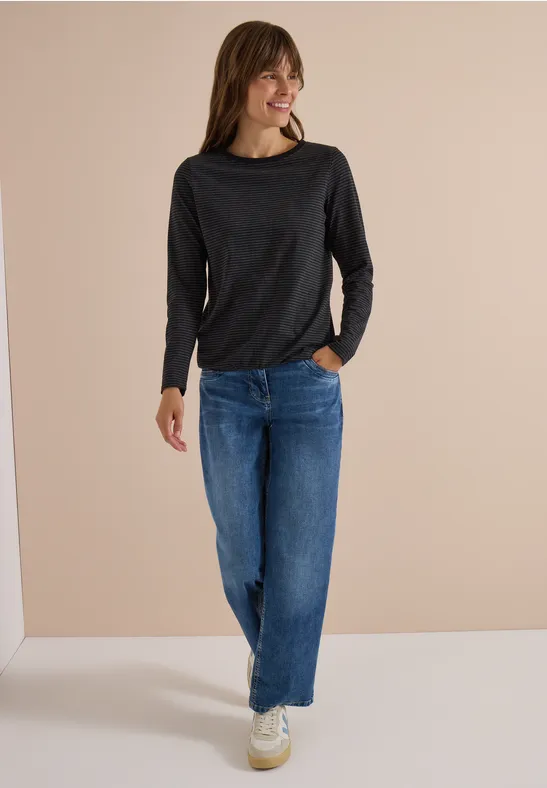 Femme souriante portant un t-shirt à manches longues noir et un jean large bleu sur fond neutre.