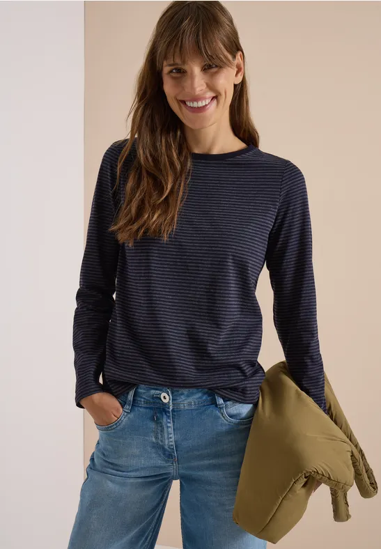 Femme en jean et pull rayé, tenant une veste, souriante.