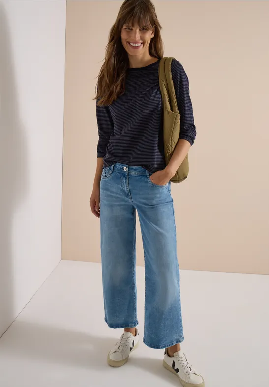 Jeune femme souriante en pull bleu marine, jean large et baskets, posant devant un fond épuré.