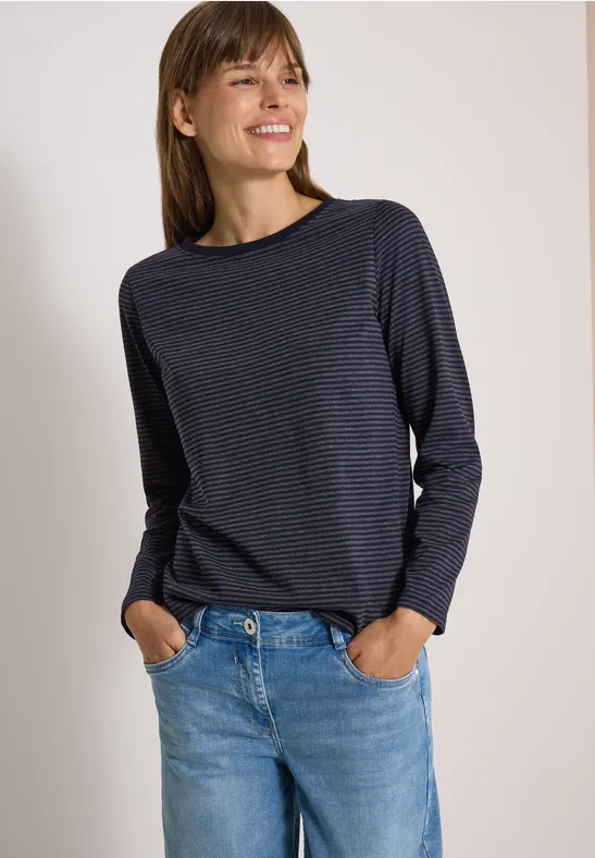 Femme en t-shirt manches longues bleu marine et jean, mains dans les poches, sourire.