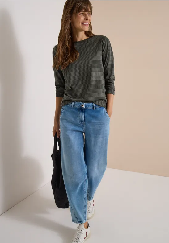 Femme en haut vert foncé à manches 3/4, jean large, baskets blanches et sac noir sur fond clair