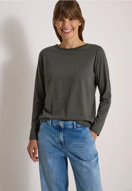 Une femme souriante porte un haut vert olive à manches longues et un jean bleu décontracté, main dans la poche.