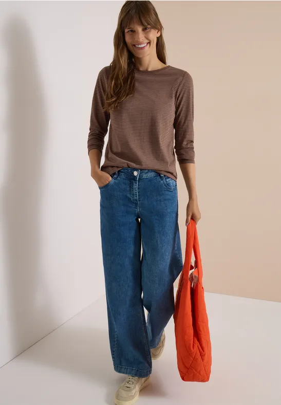 Frau in braunem Langarmshirt, weiten Jeans und weißen Sneakers hält eine orangefarbene Tasche und lächelt.