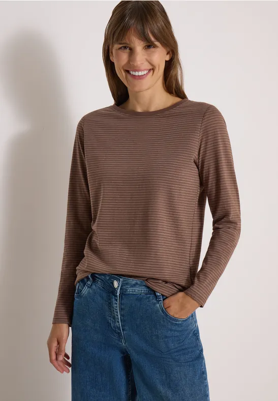 Frau in braunem gestreiftem Langarm-Shirt und blauen Jeans lächelnd vor neutralem Hintergrund.