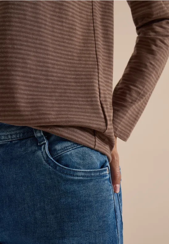 Person in braun gestreiftem Oberteil mit Hand in Jeans-Tasche vor beigem Hintergrund.