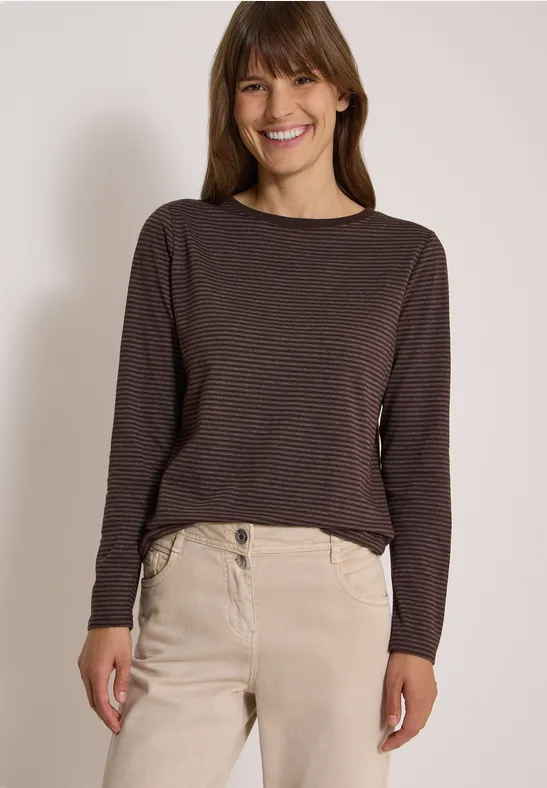 Eine lächelnde junge Frau trägt ein dunkelgestreiftes Langarmshirt und helle Hosen vor hellem Hintergrund.