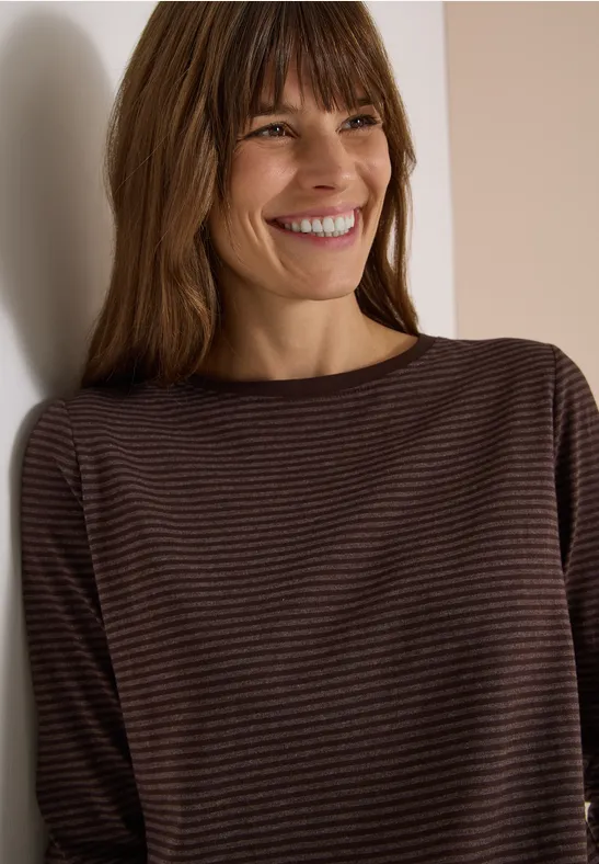 Eine lächelnde Frau mit braunen Haaren in einem gestreiften braunen Langarmshirt lehnt an einer weißen Wand.