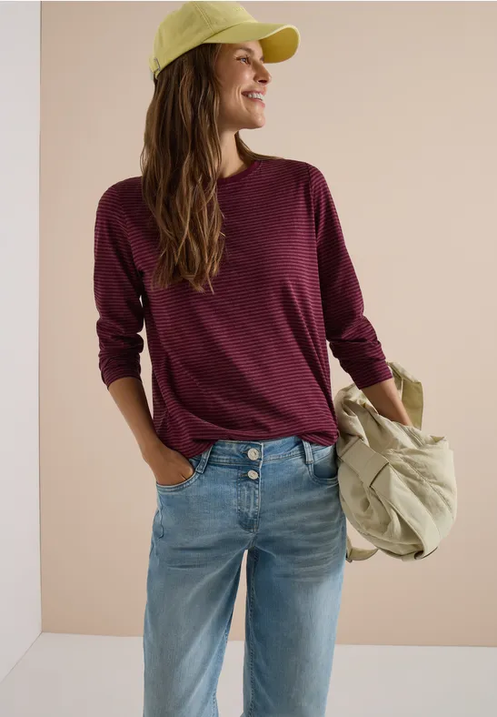 Femme en pull bordeaux, jean et casquette verte, tenant un sac sur l’épaule, souriant de profil.