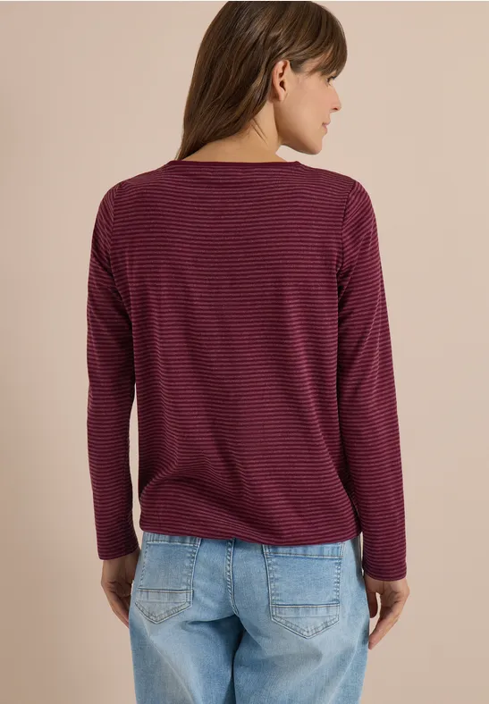 Vue de dos: femme en pull manches longues bordeaux à fines rayures et jean bleu devant un mur beige.