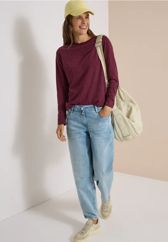 Femme en casquette jaune, pull bordeaux, jean clair et grand sac beige contre un mur clair.