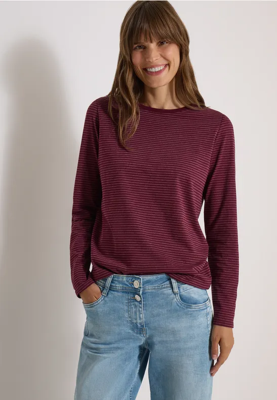 Femme en pull bordeaux à manches longues et jean, souriante, mains dans les poches.