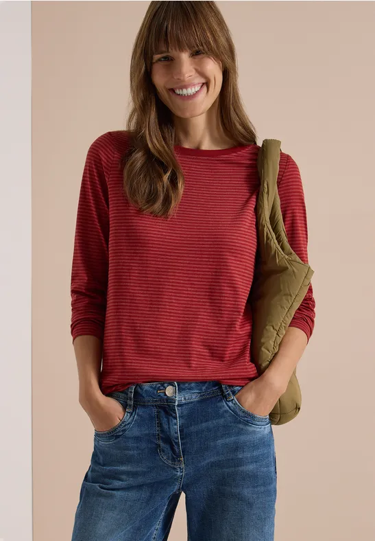 Lächelnde Frau in rotem Langarmshirt, blauen Jeans und olivgrüner Umhängetasche vor neutralem Hintergrund.