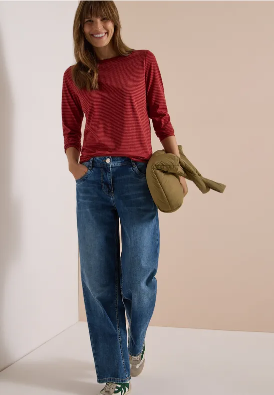 Eine junge Frau in rotem Langarmshirt und weiten Jeans hält ein grünes Cap und lächelt.