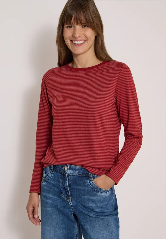 Eine lächelnde Frau trägt ein rotes, langärmliges Shirt mit feinen Streifen und blaue Jeans.