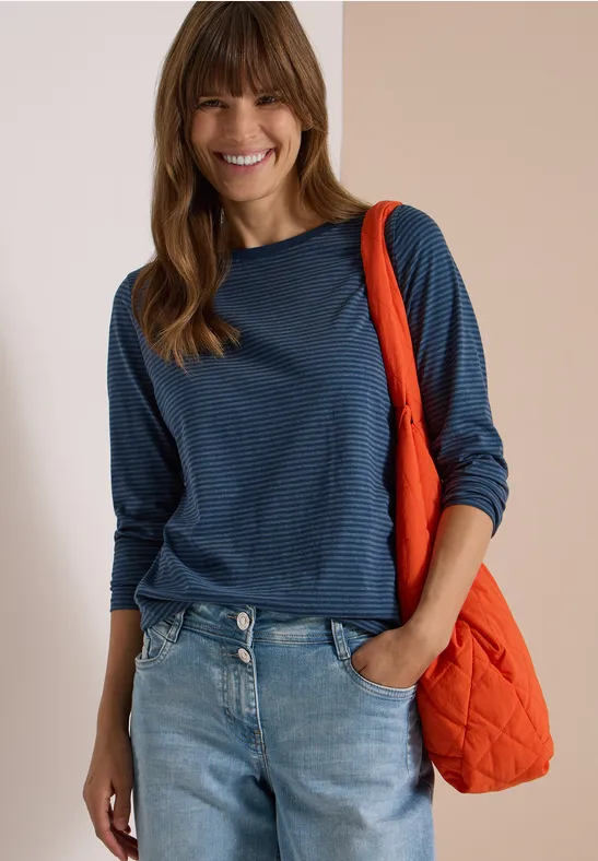 Lächelnde Frau in blau gestreiftem Langarmshirt und Jeans, mit orangefarbener Stepp-Tasche über der Schulter.