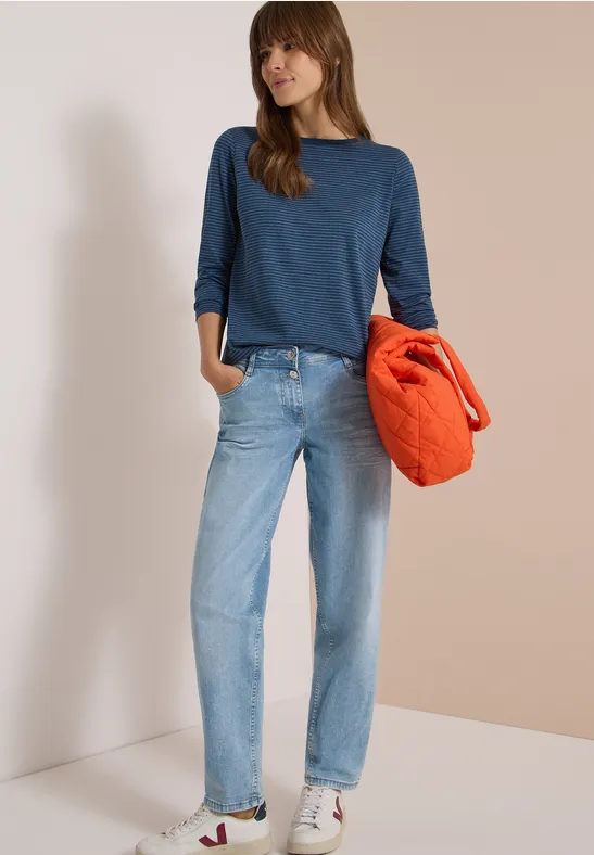 Frau in Jeans, blauem Pullover und weißen Sneakern hält eine orange Jacke vor neutralem Hintergrund.