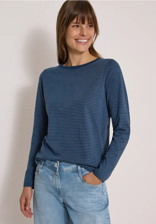 Frau mit blau gestreiftem Langarmshirt und Jeans lächelt, Hände in den Taschen