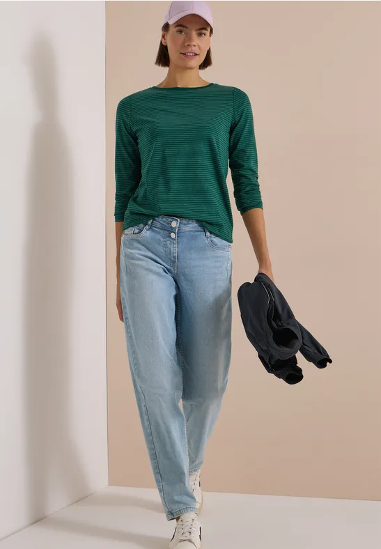 Frau in grünem Dreiviertelshirt, Jeans und Cap hält Jacke und geht vor schlichtem Hintergrund.