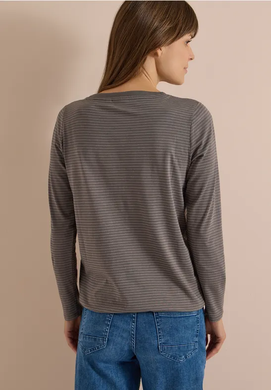 Frau von hinten, trägt graues gestreiftes Langarmshirt und blaue Jeans vor beiger Wand.