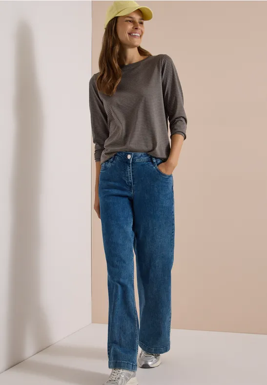 Eine Frau trägt graues Langarmshirt, blaue Jeans, weiße Sneaker und gelbe Kappe und lächelt.