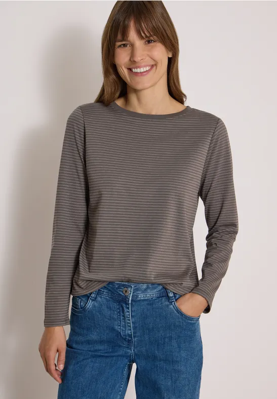 Eine lächelnde Frau trägt ein grau gestreiftes Longsleeve und blaue Jeans vor hellem Hintergrund.