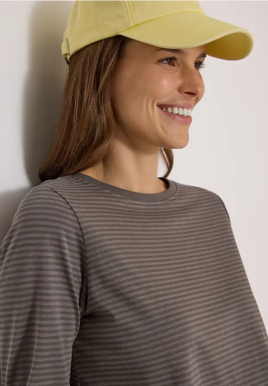 Eine Frau mit gelber Kappe lächelt, trägt ein gestreiftes Shirt und lehnt an hellem Hintergrund.