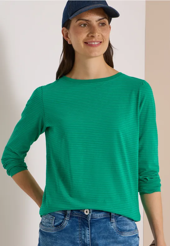 Junge Frau mit grüner Bluse und blauer Baseballkappe lächelt vor neutralem Hintergrund.