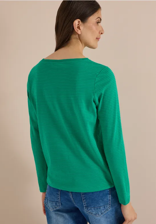 Frau von hinten mit grünem Langarmshirt und blauer Jeans vor hellem, einfarbigem Hintergrund.