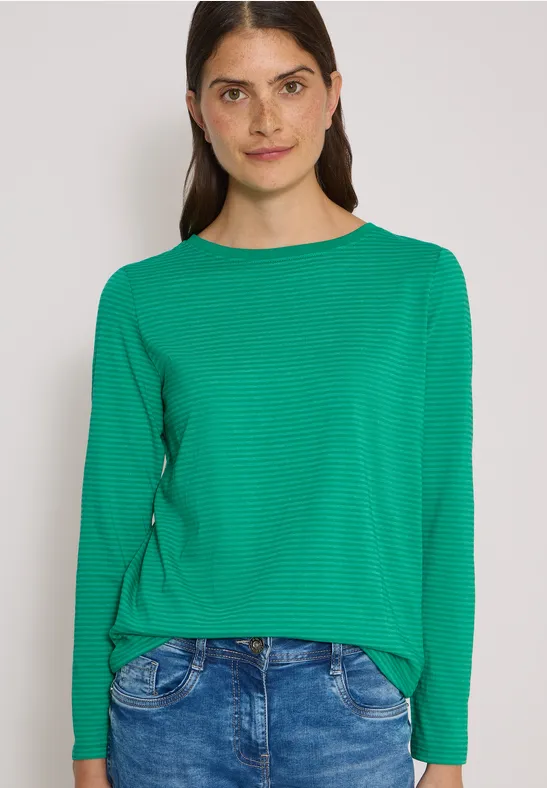 Eine Frau mit langärmligem grünen Shirt und Jeans vor weißem Hintergrund lächelnd.