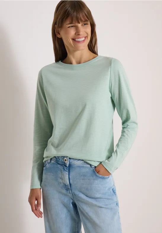 Eine lächelnde Frau trägt ein mintgrünes Langarmshirt und eine helle Jeans