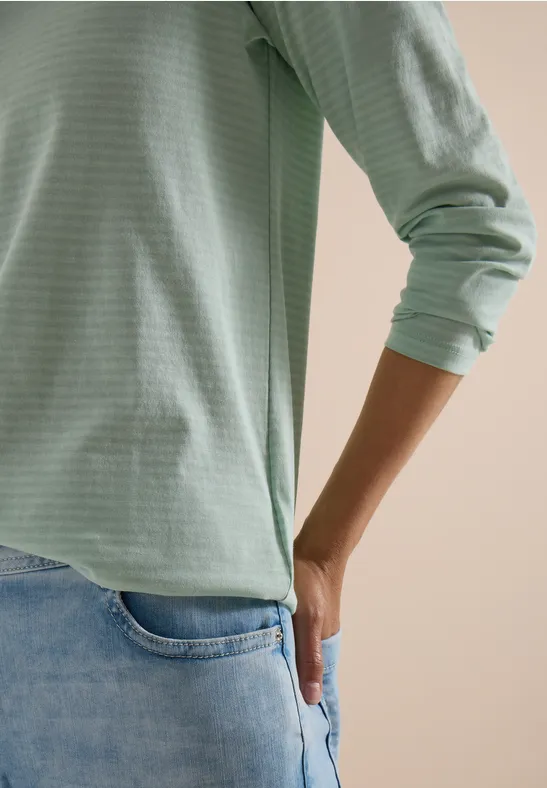 Person trägt hellgrünes Langarmshirt und Jeans, Hand steckt lässig in Gesäßtasche.