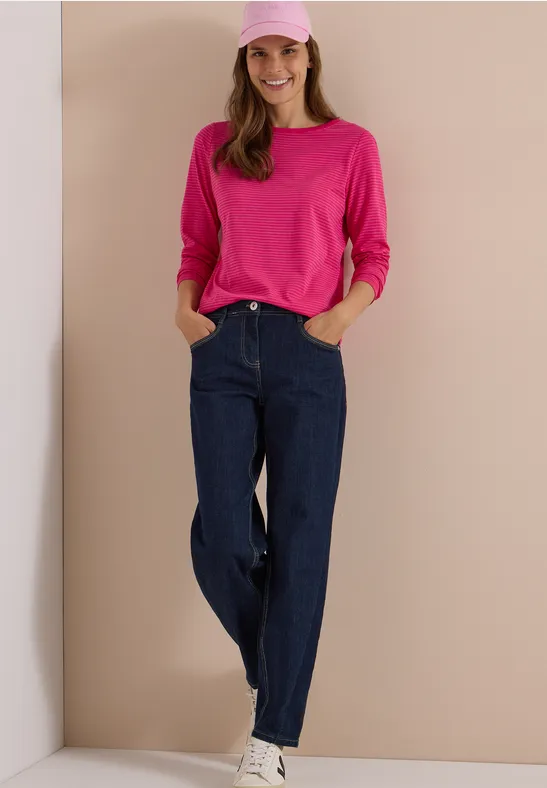 Eine Frau in dunkelblauen Jeans, pinkem Longsleeve und rosa Mütze steht vor hellem Hintergrund und lächelt.
