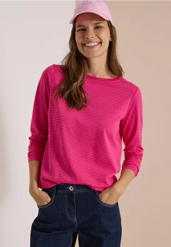 Eine lächelnde Frau mit pinkem Shirt und Cap, lässig in Jeans posierend.