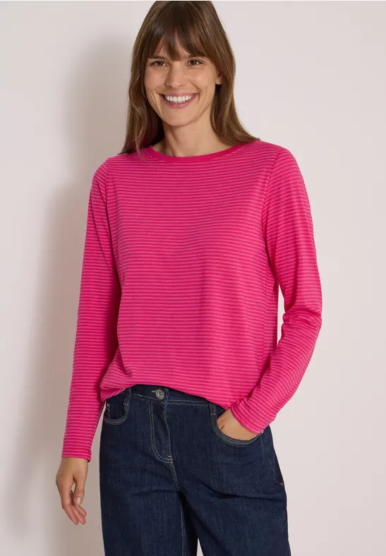 Frau in pinkem Langarmshirt und Jeans, lächelnd vor hellem Hintergrund.