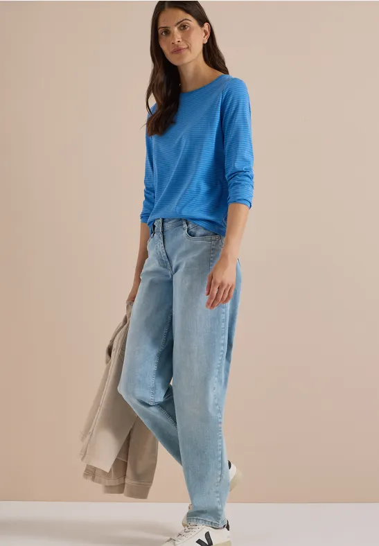 Frau in blauem Langarmshirt, weiten Jeans und Sneakers vor neutralem Hintergrund, hält helle Jacke.