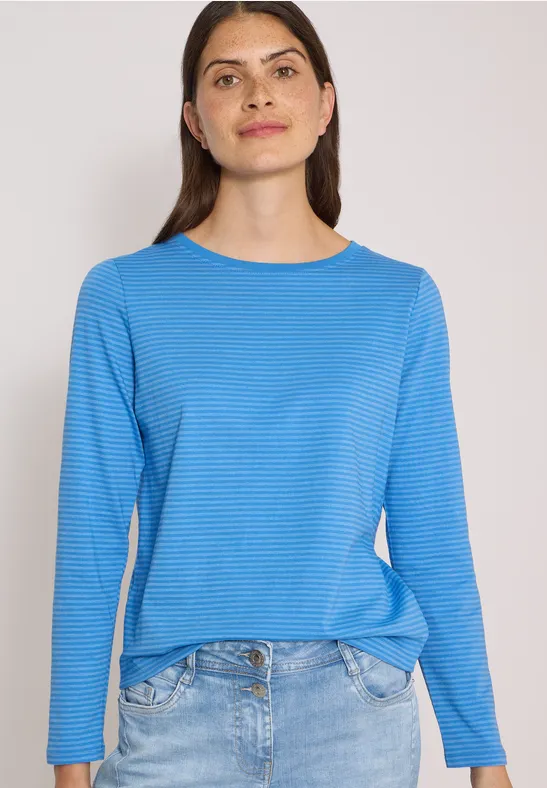 Frau in blauem Langarmshirt und Jeans vor hellem Hintergrund.